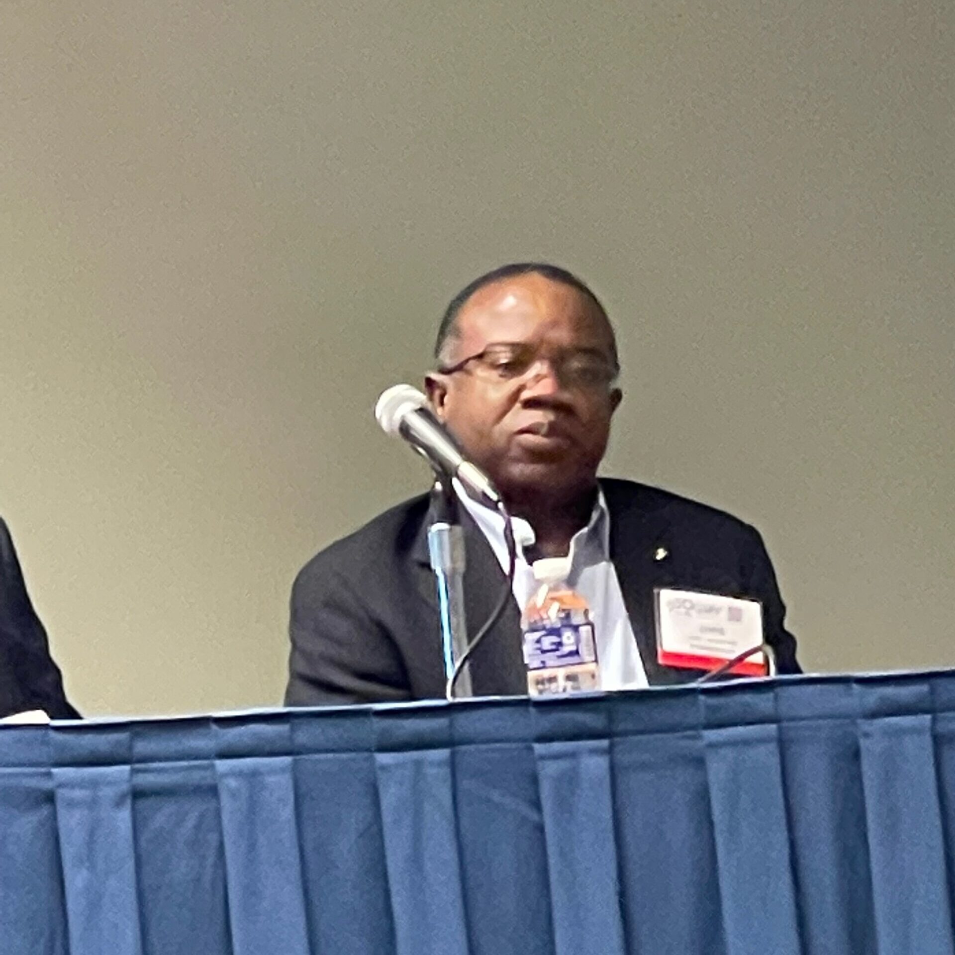 Photo of a panel discussion featuring a speaker seated at a table with a microphone. The individual is dressed in formal attire, wearing a black blazer, and appears to be actively engaged in the discussion. A bottle of water is visible on the table, and the background is plain, emphasizing the focus on the speaker.