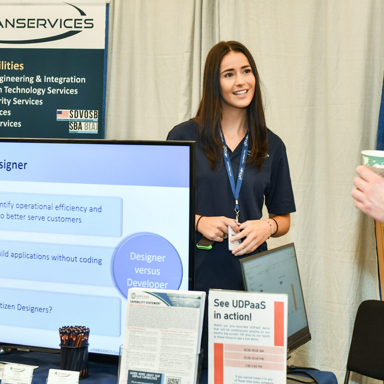 An eTRANSERVICES booth at an event, featuring a presenter speaking with a visitor. The booth showcases key capabilities, services, and products, including information about Citizen Designers and the UDPaaS platform.