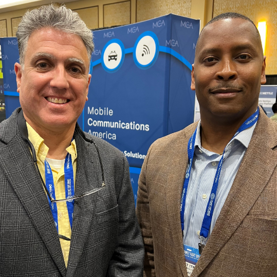 The image shows two men standing together at an event, smiling for the camera. They are standing in front of a display for Mobile Communications America (MGA). The display features a blue and white backdrop with the text "Mobile Communications America" and icons representing wireless communication solutions. Both individuals are wearing name tags and lanyards, indicating they are participants at the event.