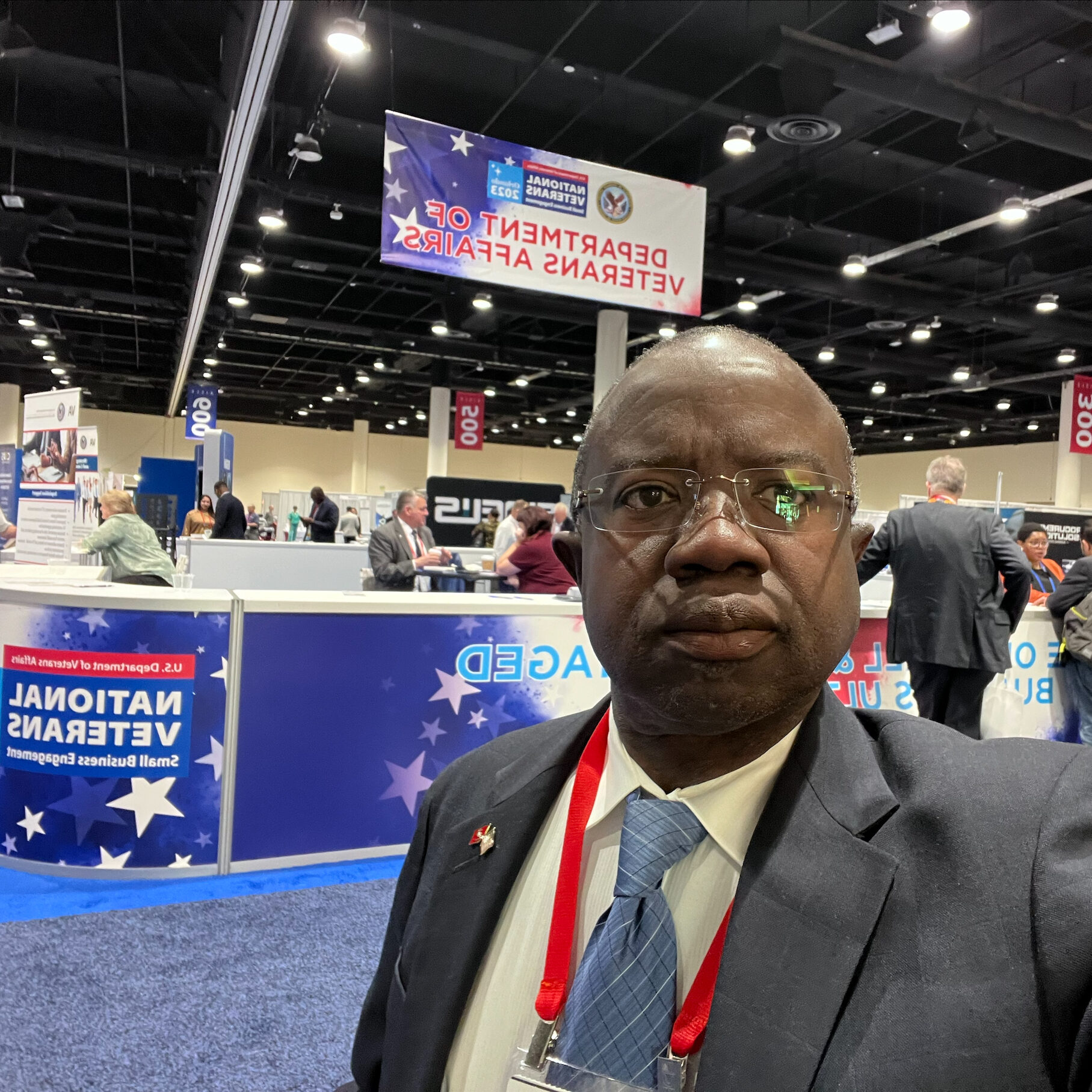 The image shows a self-portrait (selfie) taken at a conference or event. In the background, you can see the Department of Veterans Affairs booth, with a banner for National Veterans 2023 and Small Business Empowerment. The conference hall appears to have several other booths and participants, with attendees interacting and walking around. The individual in the selfie is dressed in a suit and has a name tag and lanyard on.