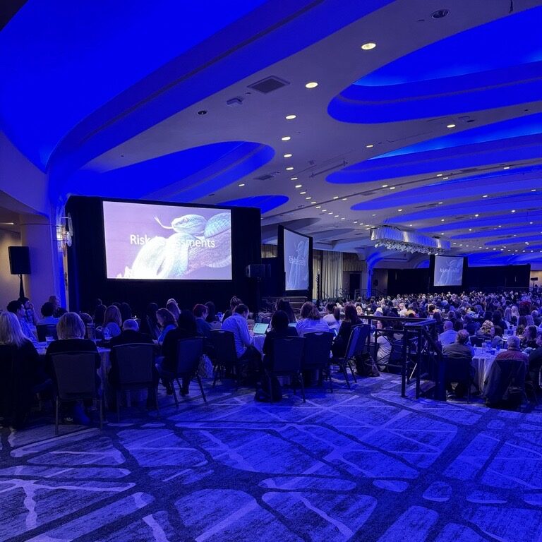The image shows a large conference room filled with attendees. The room is lit with blue lighting, creating a vibrant atmosphere. In the front of the room, there is a large screen displaying the words "Risk Elements," indicating that the session might be focused on risk management or similar topics. The attendees are seated at tables, some appearing to take notes or engage with the presentation. The room has a modern design with curved ceilings and a stylish carpet pattern.