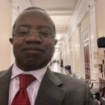 The image shows a selfie of a person standing in a hallway. The individual is wearing a gray suit jacket with a red tie and glasses. The hallway appears to be an elegant, well-lit area with ornate ceiling fixtures and classic architectural details, including columns and doors along the walls. The person appears to be smiling slightly, and there is a sense of a formal or professional setting in the background.