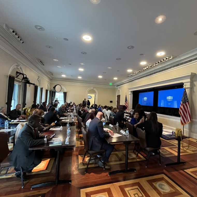 The image shows a meeting room with attendees seated at a long table. The room is decorated with American flags, and two screens on the wall display the White House logo. The participants appear to be engaged in a discussion or listening to a presentation. The atmosphere seems formal, with individuals dressed in business attire. The setting likely represents an important government or professional event.