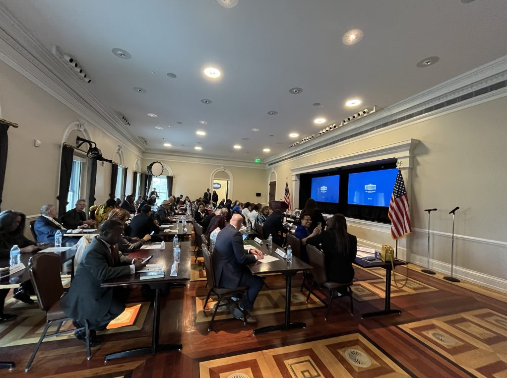 Screenshot 2024-03-18 154147 The image shows a meeting room with attendees seated at a long table. The room is decorated with American flags, and two screens on the wall display the White House logo. The participants appear to be engaged in a discussion or listening to a presentation. The atmosphere seems formal, with individuals dressed in business attire. The setting likely represents an important government or professional event.