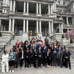 The image shows a large group photo of individuals standing together on the steps of a grand building. The group is diverse, with both men and women dressed in formal attire. Some people are smiling, while others stand with a more serious expression. The setting appears to be a formal or professional event, with elegant architecture in the background.