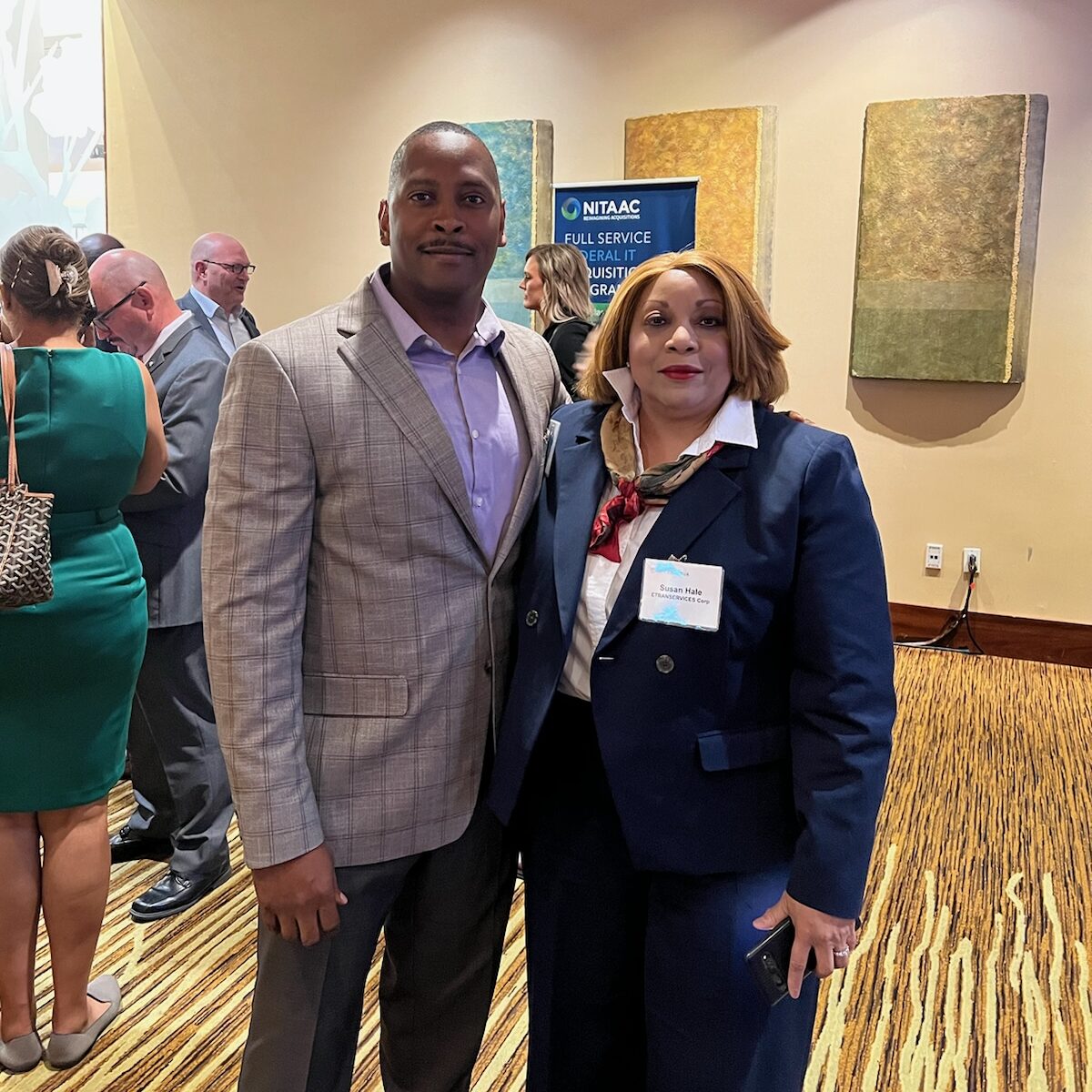The image shows two individuals posing together at an event. The man is dressed in a light gray suit, and the woman is wearing a navy blue blazer with a red scarf and a name tag that reads "Susan Hale." The background shows a banner for NITAAC (National Institutes of Health Information Technology Acquisition and Assessment Center) and a crowd of people mingling at the event. The setting appears to be a professional networking event.