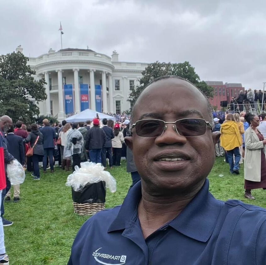 The image shows a person taking a selfie in front of the White House during an event. The individual is wearing a blue eTRANSERVICES branded polo shirt, and there are many people in the background, some of whom are dressed in rain gear. The event appears to be outdoors with cloudy weather, and there is a stage setup in front of the White House, suggesting a public gathering or speech.