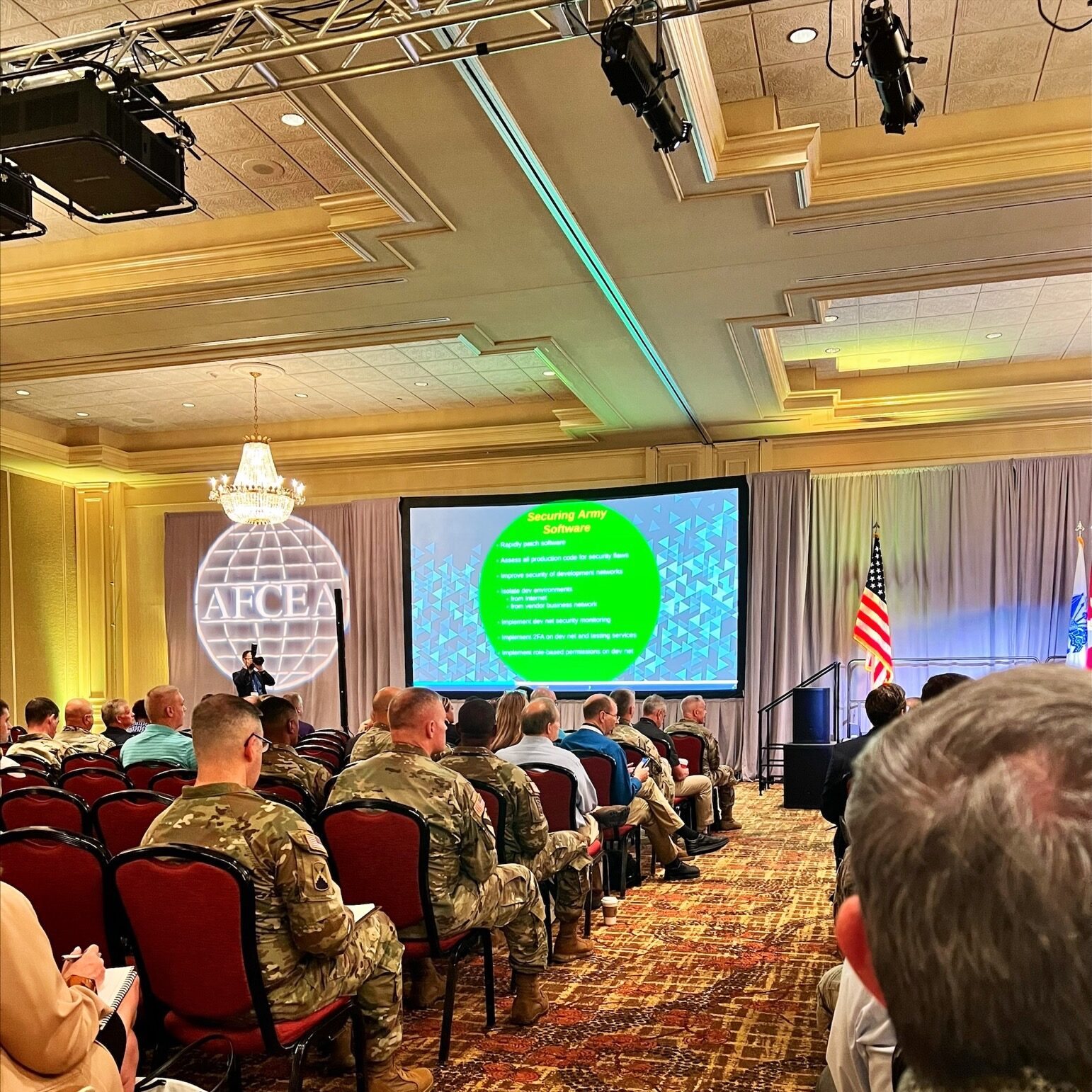MicrosoftTeams-image (2) The image shows a conference or seminar setting with attendees seated in rows, listening to a presentation. Some of the audience members are in military attire, and the speaker is presenting a topic titled "Securing Army Software" on a large screen. The room has a formal atmosphere, likely at an AFCEA event, with U.S. flags displayed in the background. The audience appears focused on the presentation.