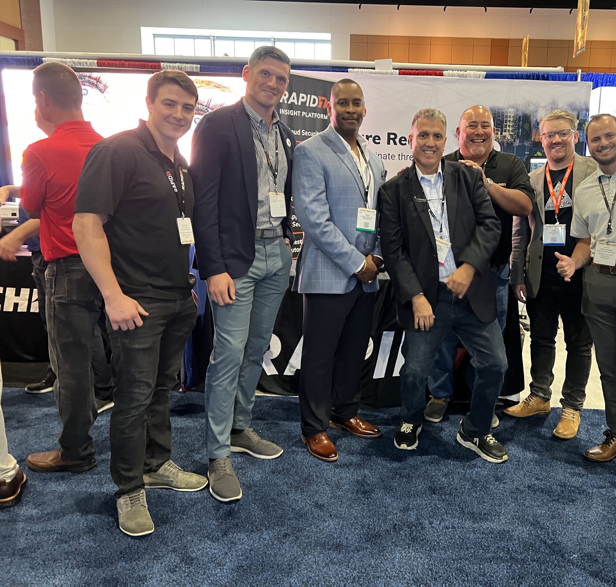 MicrosoftTeams-image (4) The image shows a group of six people posing together at an event or conference. They are standing in front of a booth with a banner featuring the Rapid7 logo. The individuals are dressed in a mix of business and casual attire, smiling and engaging with each other. The setting appears to be an exhibition hall, and the group is likely involved in promoting products or services related to Rapid7.