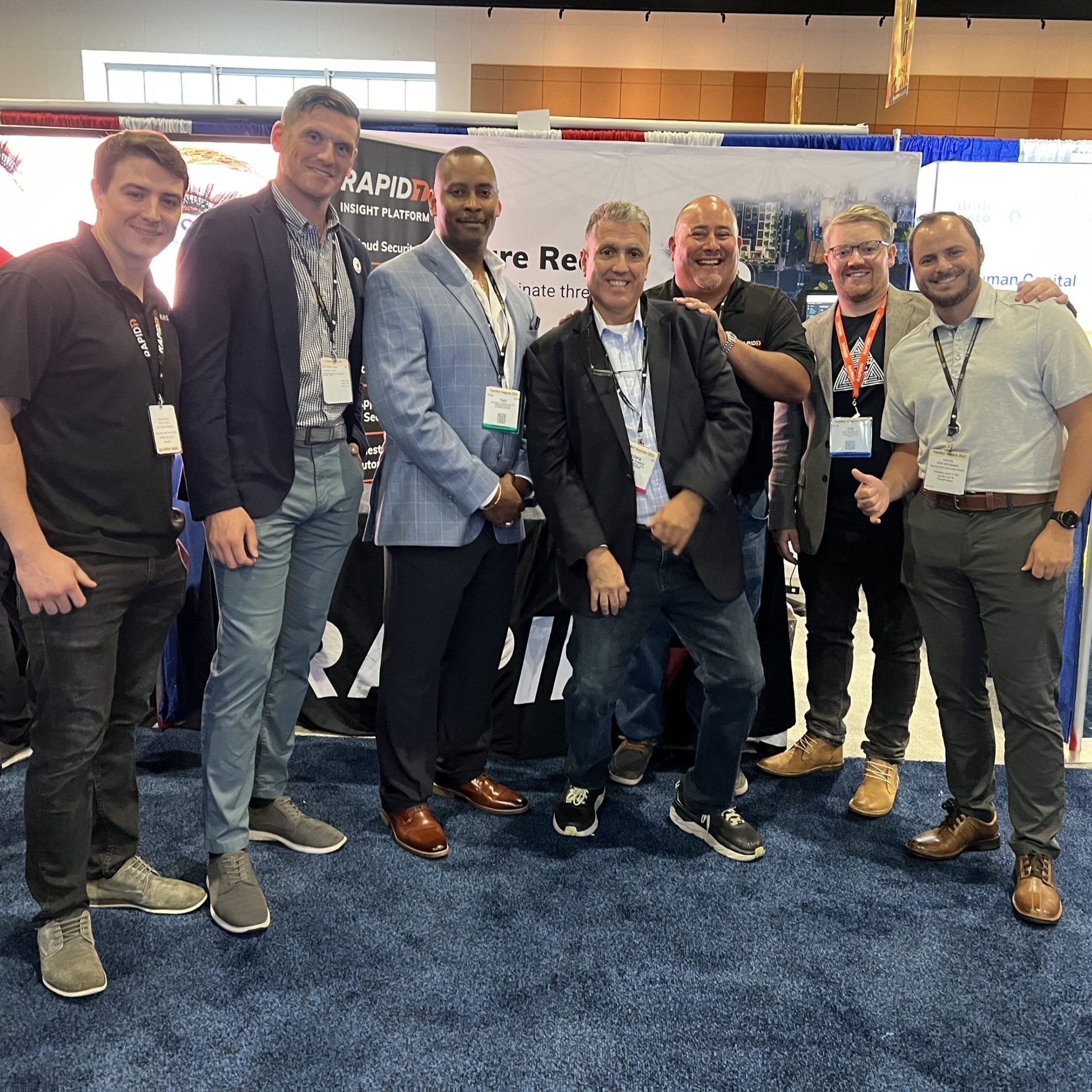 The image shows a group of six people posing together at an event or conference. They are standing in front of a booth with a banner featuring the Rapid7 logo. The individuals are dressed in a mix of business and casual attire, smiling and engaging with each other. The setting appears to be an exhibition hall, and the group is likely involved in promoting products or services related to Rapid7.